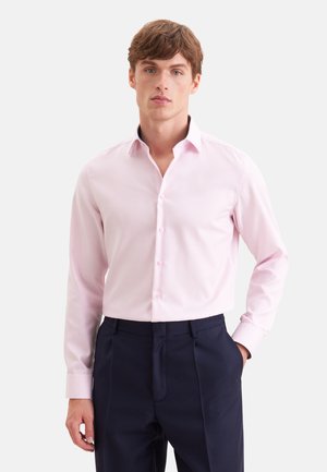 Jeune homme portant une chemise rose clair à boutons et un pantalon bleu marine foncé, debout avec une main dans la poche sur un fond uni.