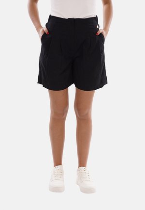 Donna che indossa pantaloncini neri a vita alta con tasche laterali, scarpe da ginnastica bianche e top bianco, in piedi con le mani nelle tasche.