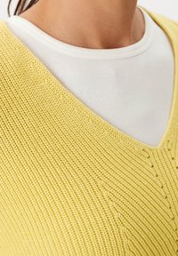Gelber Strickpullover mit V-Ausschnitt-Design, das ein strukturiertes Muster aufweist, über einem weißen, gerippten Shirt mit rundem Ausschnitt getragen.