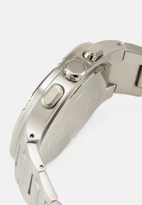 Sølv rustfritt stålarmbåndsur med strukturert overflate, fremtredende knapper og en jevn, polert bezel, med et intrikat armbånddesign.