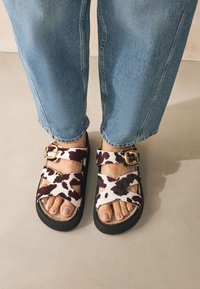 Lehmamustriga sandaalid pehme, tekstureeritud pealmise osaga ja reguleeritavate pandlatega. Paksud mustad taldmikud tagavad stabiilsuse. Kandmisega hele sinine teksariie.