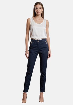 Donna in piedi che indossa una camicetta bianca senza maniche, pantaloni blu navy con bottoni decorativi e scarpe con tacco color nude su sfondo bianco.