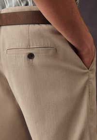 Pantalones beige con una tela texturizada, que presentan un bolsillo trasero con un botón marrón y un cinturón marrón con un patrón.
