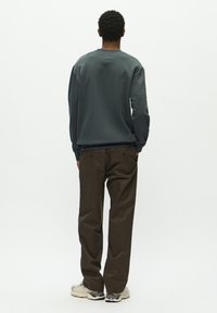 Dunkelgrauer Rundhals-Sweatshirt, kombiniert mit braunen Hosen. Die Sneakers haben ein grau-schwarzes Design. Insgesamt glatte Textur.