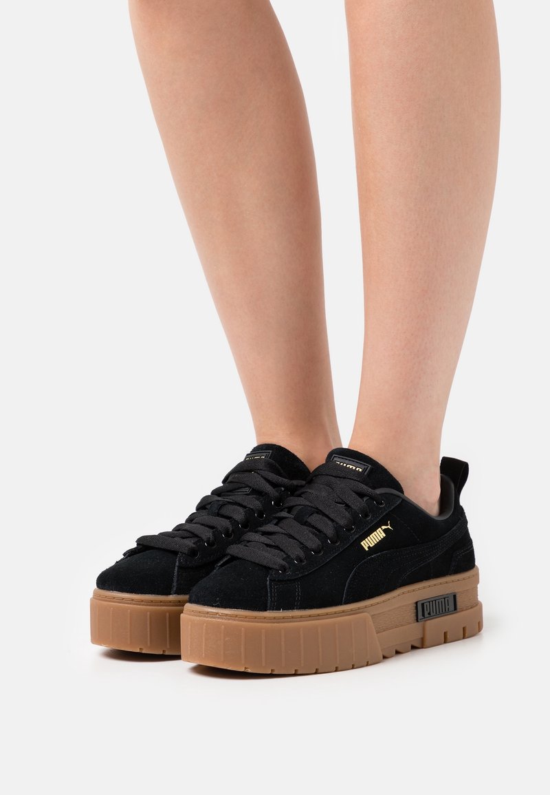 Baskets noires Puma avec semelles épaisses marron portées sur une paire de jambes nues, montrées sur un fond blanc uni.