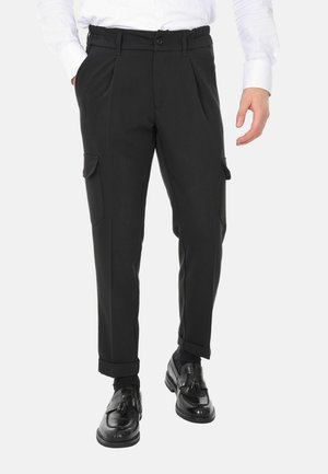 Uomo che indossa pantaloni cargo neri con risvolti e tasche con patta, mocassini neri con nappine e camicia bianca a maniche lunghe, in piedi su sfondo bianco.