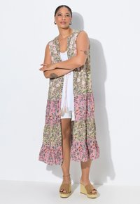 Gilet floral long en motifs beiges, roses et verts avec des bords à volants et une taille cintrée. Associé à des shorts blancs et des sandales à plateforme.