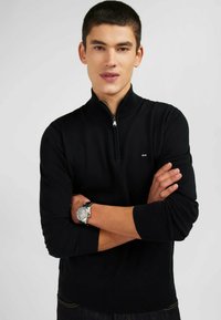 Pull à col zippé noir en tissu maille doux, avec des manches longues, des poignets et un ourlet côtelés, et un petit logo sur la poitrine.