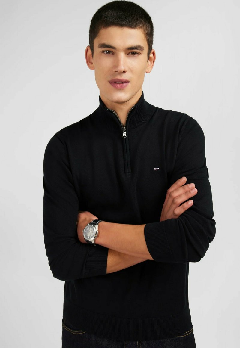 Pull à col zippé noir en tissu maille doux, avec des manches longues, des poignets et un ourlet côtelés, et un petit logo sur la poitrine.