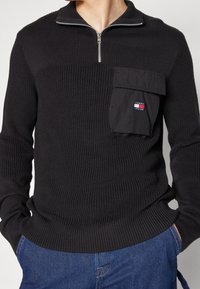 Schwarzer Strickpullover mit einem Reißverschlusskragen, ausgestattet mit einer Brusttasche und einem kleinen Logo-Patch. Gerippte Textur und taillierte Passform.