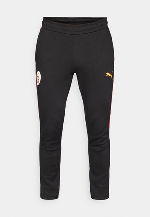 Svart joggebukser med røde sideakenter, med en hvit logo-lapp og en gul Puma-logo. Laget av mykt, elastisk stoff.