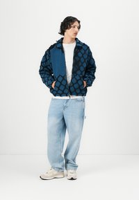 Blue patterned jacket with black geometric design, fleece texture. Paired with light blue jeans and beige sneakers. Simple white t-shirt underneath.