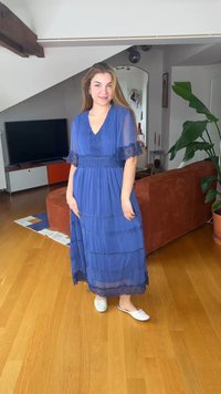 Jonge vrouw in een blauwe jurk met kanten rand en witte slippers, staand op een houten vloer in een modern woonvertrek.