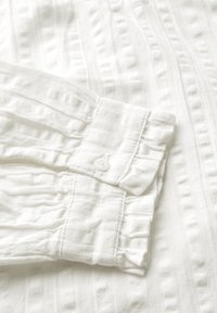 Camicia bianca in cotone con tessuto testurizzato e sottili strisce, polsini con bottoni e dettagli arricciati lungo le maniche.