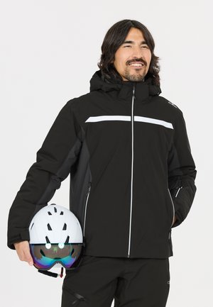 Mand iført en sort skijakke, der holder en hvid skihjelm med reflekterende goggles, smilende og kigger lidt til siden.