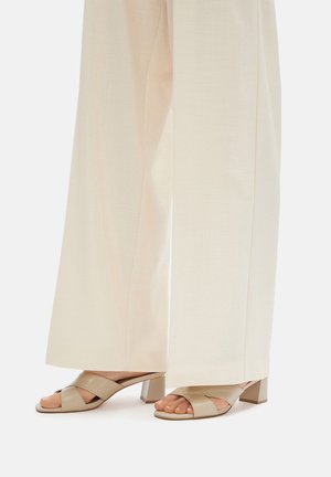 Beżowe sandały na obcasie z krzyżowym paskiem, w zestawieniu z jasnymi, szerokimi spodniami. Widoczna gładka tekstura i smukły blokowy obcas.