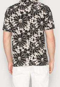 Camicia a maniche corte nera e beige con un motivo floreale. Presenta grandi margherite e fiori più piccoli. Tessuto morbido, vestibilità comoda, con colletto.