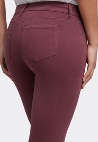 Pantalones de algodón burdeos con textura suave, cintura alta y dos bolsillos traseros con un diseño en forma de diamante. Vista cercana desde el lado.