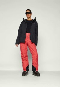 Schwarze, isolierte Jacke mit Kapuze, kombiniert mit roten Skihosen mit dunkelblauen Akzenten. Das Modell trägt schwarze Skischuhe und Sonnenbrille.