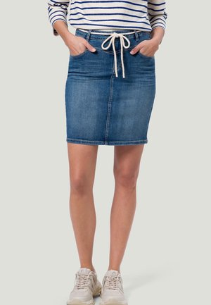Woman wearing a knee-length blue denim skirt with a white rope belt, striped long-sleeve shirt, and white sneakers, standing with hands in pockets.