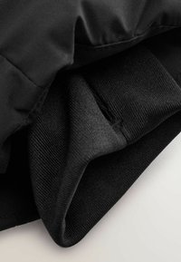Schwarze gepolsterte Jacke mit einem glatten Außenstoff und strukturierten Bündchen. Eine Nahaufnahme zeigt das Innenfutter und die Nähdetails.