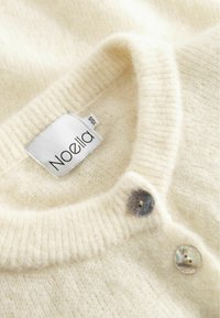 Kremkrāsas adīta jaka ar ribotu apkakli, divām dekoratīvām pelēkām pogām un piešūtu birku ar uzrakstu "Noella". Mīksta, pūkaina tekstūra.