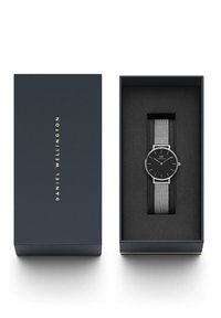 Montre-bracelet avec bracelet en maille argentée et cadran noir sur un coussin en mousse noire à l'intérieur d'une boîte sombre étiquetée Daniel Wellington.