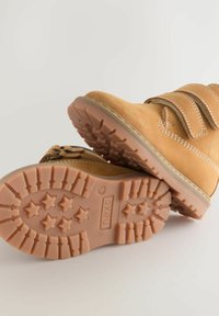 Beige Wildlederstiefel mit Klettverschluss, ausgestattet mit einer Gummisohle mit Sternmustern und einer gestickten Akzentlinie entlang der oberen Kante.