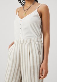 Top bianco senza maniche con bottoni e scollo a V, abbinato a pantaloni a vita alta beige e bianchi con righe e tasche laterali.