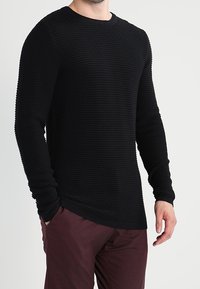 Pull noir à manches longues avec un design en côtes texturées, col rond et coupe ajustée, associé à un pantalon bordeaux.