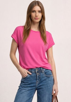 Kobieta z długimi brązowymi włosami, ubrana w różowy t-shirt z krótkim rękawem i niebieskie dżinsy, pozująca z jedną ręką w kieszeni i trzymająca brązową torbę.