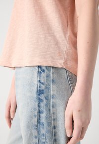 Gros plan d'une personne portant un jean bleu clair et un haut à manches courtes couleur pêche, montrant une partie du bras et de la main.