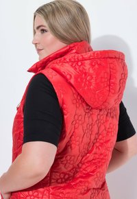 Frau in schwarzem kurzärmligem Shirt, die eine rote gesteppte Weste mit floralen Stickereien und Kapuze trägt, von der Seite und von hinten gezeigt.