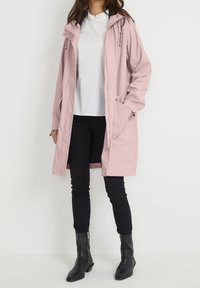 Lichtroze waterdichte parka met een capuchon, steekzakken aan de voorkant, gouden details en een midilange pasvorm. Draagt over een wit topje en zwarte broek.