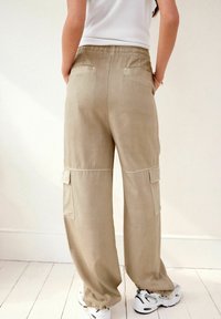 Beige cargo bukser med en afslappet pasform, elastisk talje, sidelommer og syede detaljer, vist fra ryggen med hvide sneakers.