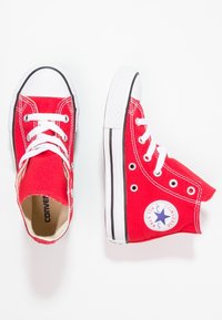 Červené plátěné vysoké tenisky mají bílé tkaničky, černou gumovou podrážku a kulaté logo Converse na kotníku. Pozadí s plochým povrchem.