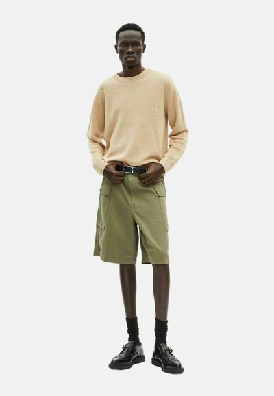 Leicht pfirsichfarbener Strickpullover, olivgrüne Cargo-Shorts mit Seitentaschen, schwarzer Gürtel, schwarze Socken und schwarze Schuhe mit perforierten Details.