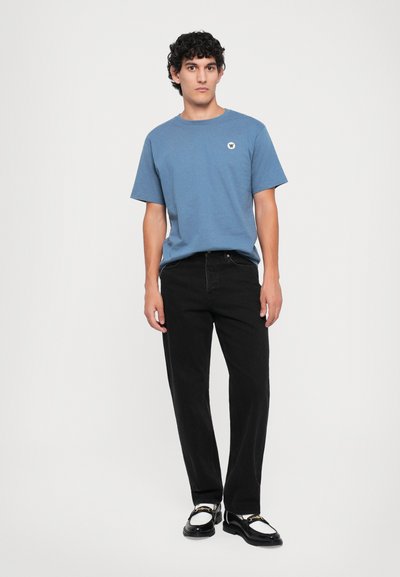 Blaues Baumwoll-T-Shirt mit Rundhalsausschnitt und kleinem weißen Logo, kombiniert mit schwarzen Jeans im Straight-Leg-Schnitt und schwarz-weißen Schuhen.