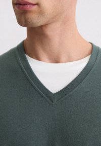 Dunkelgrüner V-Ausschnitt-Pullover aus gestricktem Material, über einem weißen Hemd getragen. Zeigt strukturierte Stoffe und ein sauberes Ausschnittdesign.