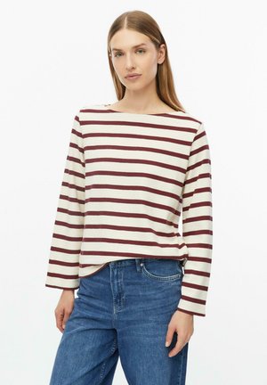 Donna con capelli lisci castano chiaro che indossa una maglia bianca a maniche lunghe con strisce orizzontali color granata e jeans blu su uno sfondo semplice.