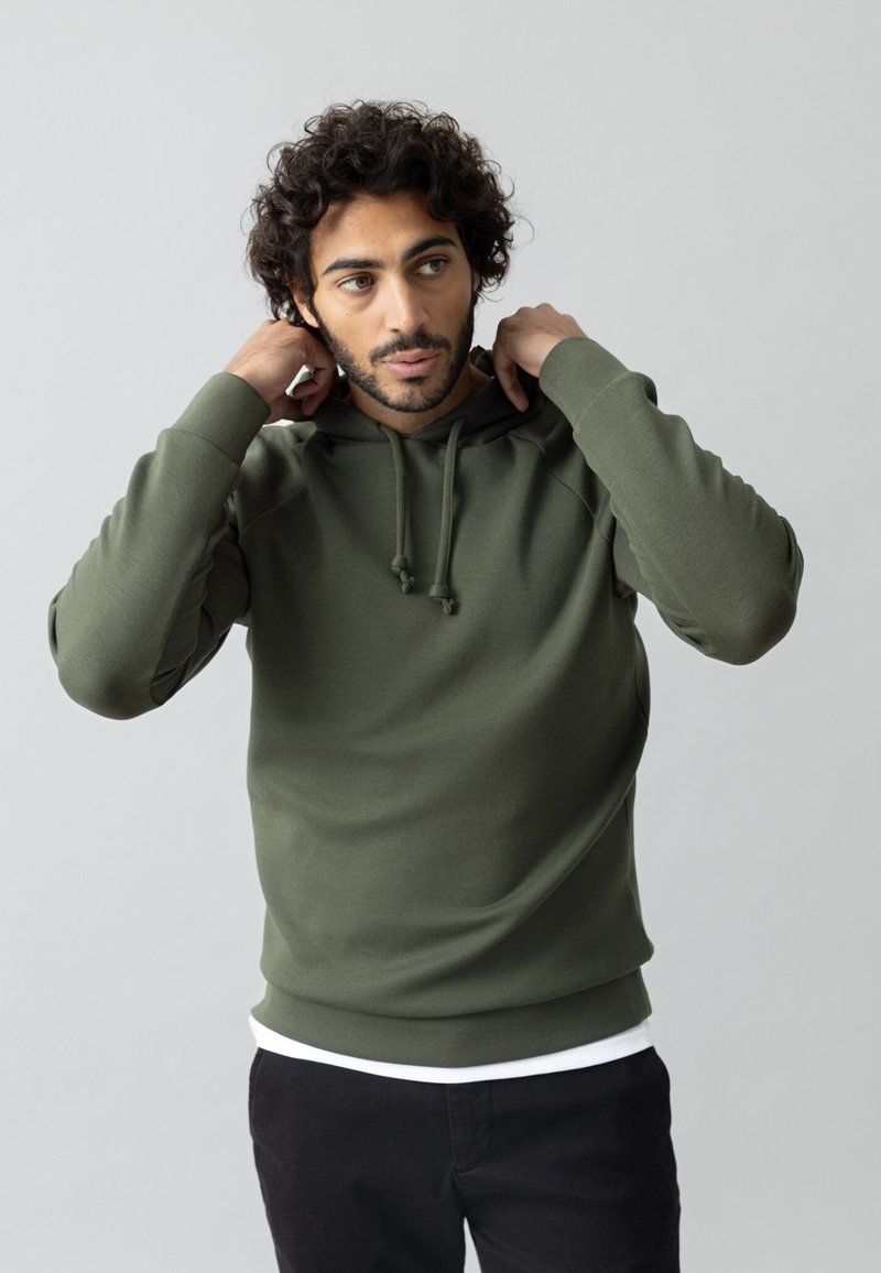 Olive roheline kapuutsiga särk, reguleeritav nöör, soonikuga varrukad ja hem, pehme tekstuur, kantud valge särgi peal, koos mustade pükstega, neutraalne taust.