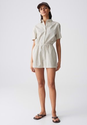 Donna che indossa una tuta corta beige a righe con coulisse in vita, cappello marrone e infradito nere, in piedi davanti a uno sfondo semplice.