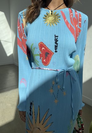 Femme portant une robe côtelée bleue avec des motifs colorés de soleil, cœur et constellation, nouée à la taille, et un grand collier en forme de soleil en or.