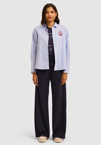 Camicia azzurra a righe con bottoni e un emblema sul petto, abbinata a pantaloni navy a gamba larga e scarpe da ginnastica beige. Tessuto morbido, vestibilità casual.
