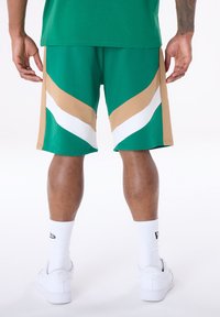 Πράσινο αθλητικό σορτς με μπεζ και λευκές λοξές ρίγες, κατασκευασμένο από ελαφρύ ύφασμα. Φοριέται με λευκές κάλτσες και αθλητικά παπούτσια.