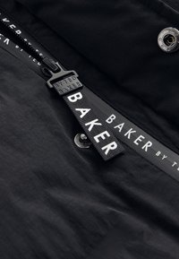 Svart tyg med en mjuk textur har en dragkedja med en dragflik som är märkt "BYTED BAKER", och en etikett som visar "BAKER by Ted". Silverfärgad knappdetalj.
