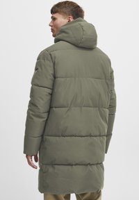Olivengrønn puffyjakke med hette, quiltet tekstur og lang lengde. Inkluderer sidelommer og minimalistiske designdetaljer.