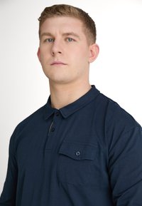 Granatowa, długorękawowa koszula polo z kołnierzykiem, trzema guzikami oraz kieszenią na piersi z guzikiem. Wykonana z miękkiego materiału, o luźnym kroju.