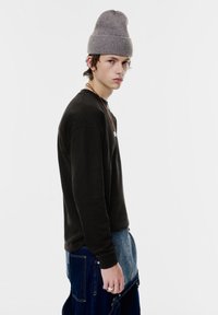 Schwarzes Langarmshirt kombiniert mit einer grauen Strickmütze; Denim-Trägerhose mit kontrastierenden blauen und grauen Einsätzen. Schlichtes Design, bequeme Passform.