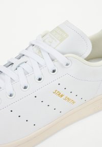 Sapatilha de couro branca com texto em dourado "STAN SMITH", detalhes perfurados e atacadores brancos planos. Textura suave e sola em tom creme.